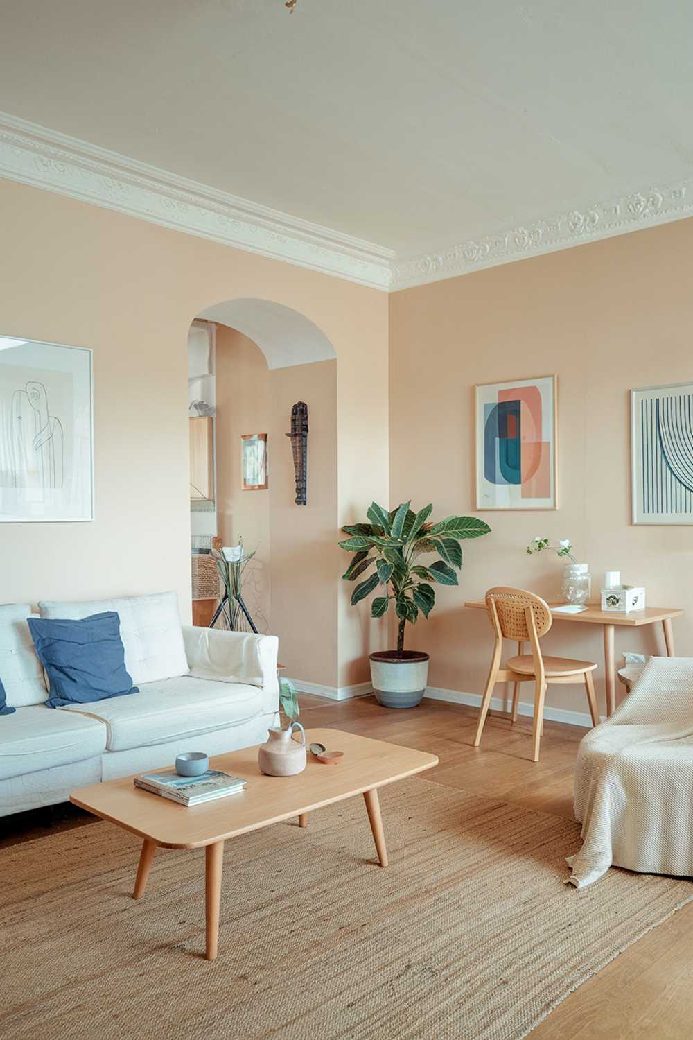 A Scandinavian-style living room in an apartment. The room features a white sofa with blue cushions, a wooden coffee table, and a beige rug. There's a potted plant in the corner. The walls are painted in soft beige, and the room has a few pieces of decorative art.