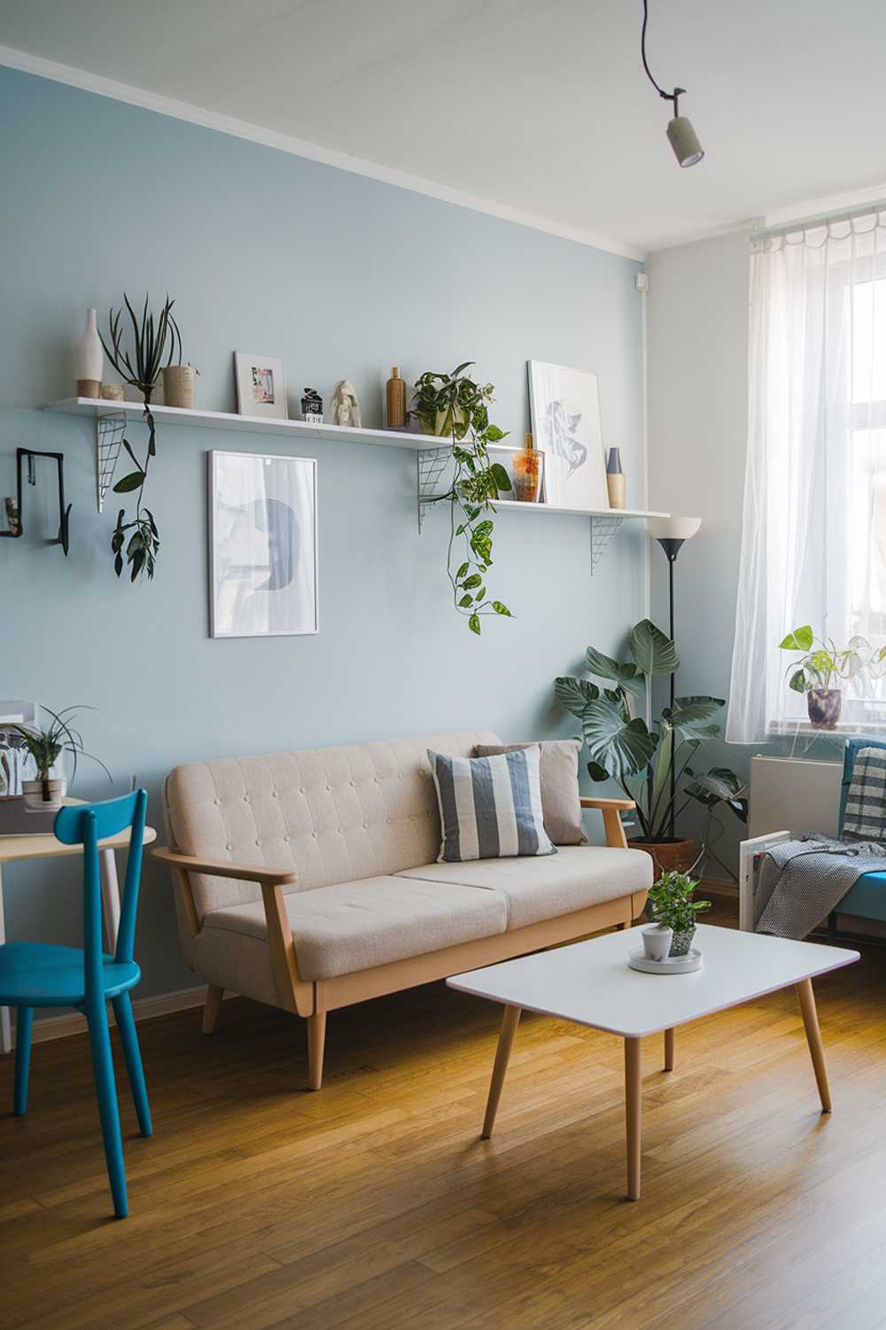 A Scandinavian-style small apartment living room. There is a beige sofa with wooden legs, a white coffee table, and a blue chair. The walls are painted in light blue. There are plants, a lamp, and decorative items on the shelves. The flooring is wooden.