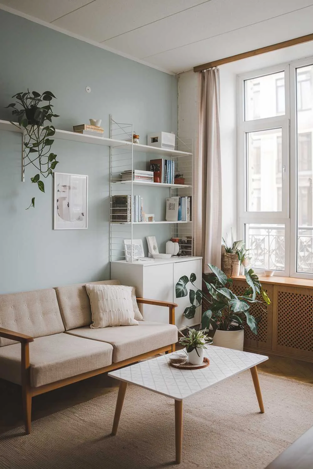 A small Scandinavian-style living room in an apartment. The room has a beige sofa with a wooden frame, a white coffee table with a geometric pattern, and a plant. There's a white bookshelf with a few books and decor items. The walls are painted in a light blue shade, and the room has a large window with a curtain. The floor is covered with a beige rug. There's a wooden panel near the window. The overall design is minimalistic and cozy.