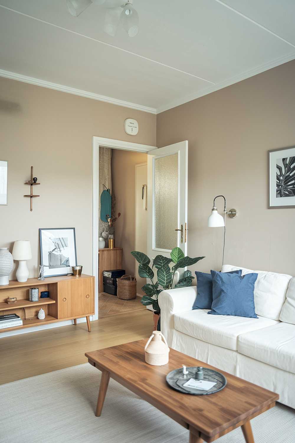 A Scandinavian style living room in an apartment. The room has a white sofa with blue cushions, a wooden coffee table, and a potted plant. There are also a few decorative items, including a lamp, a vase, and a photo frame. The walls are painted beige, and there's a door leading to another room. The flooring is a light wood.