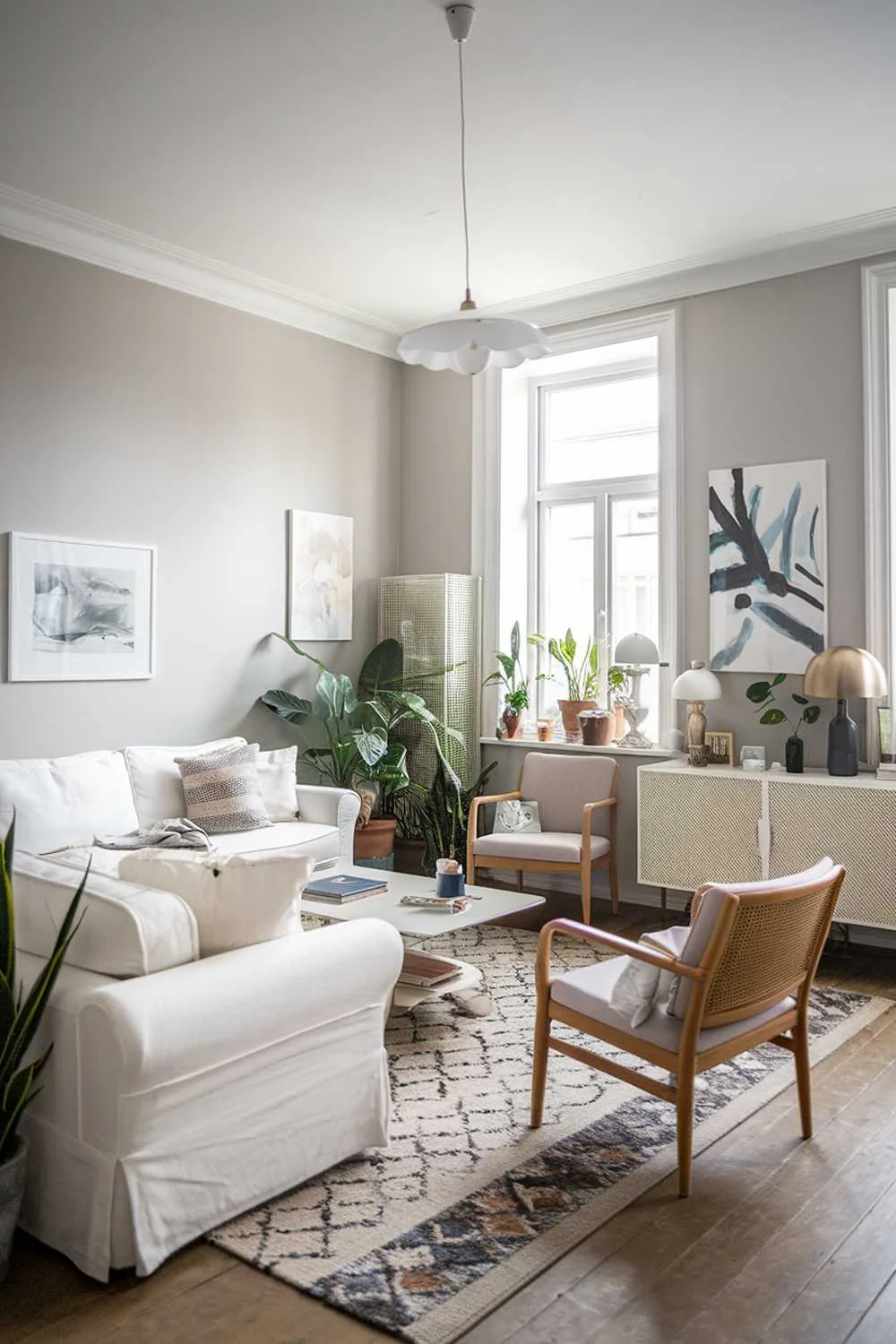 A Scandinavian-style living room with a white sofa, a few chairs, a coffee table, and a TV unit. The walls are painted in a light gray shade, and there are a few potted plants scattered around. A patterned rug is placed under the furniture. The room has a few decorative items, including a lamp, a few books, and a painting.