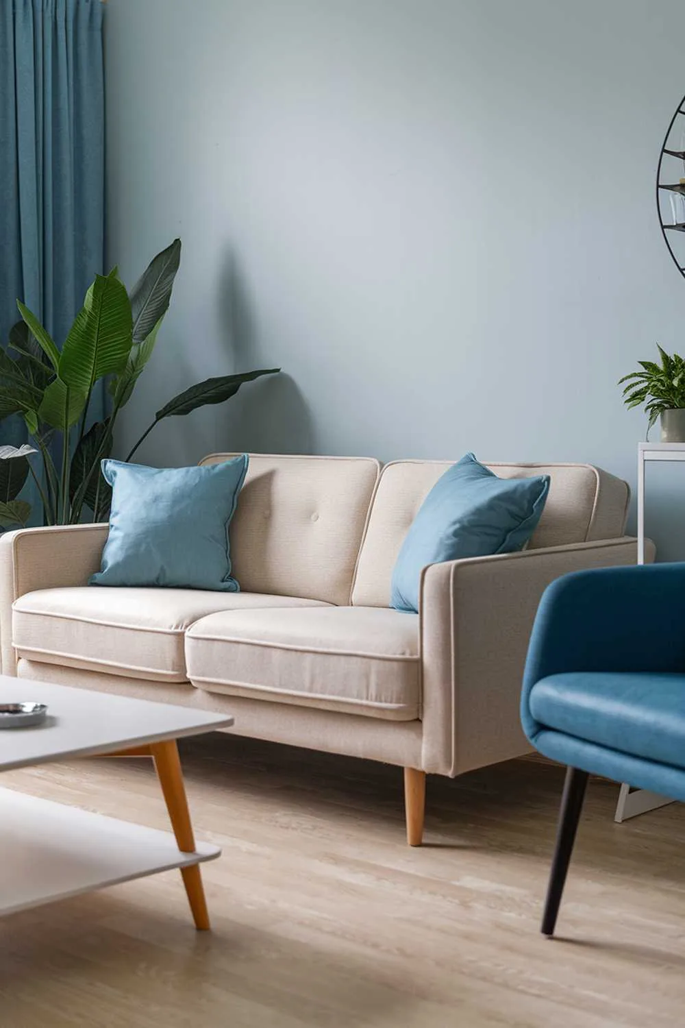 A home decor in a Scandinavian apartment living room. There is a beige sofa with blue pillows, a white coffee table, and a blue chair. The walls are painted in light blue. There is a green plant near the sofa. The floor is made of light wood.