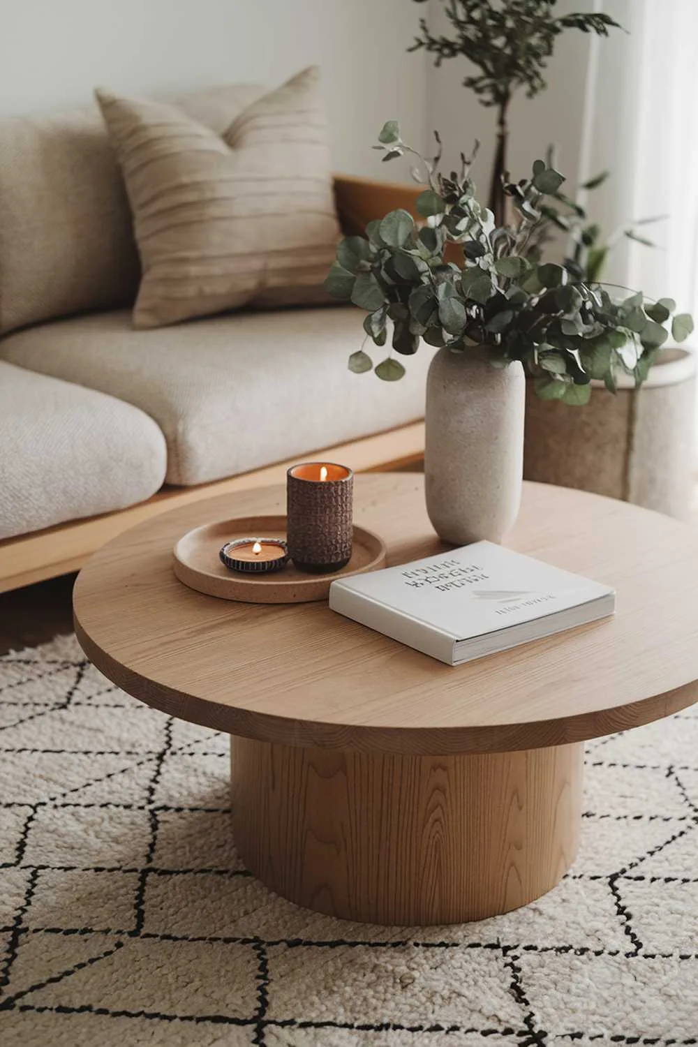 A modern Scandinavian boho living room decor. There's a beige rug with a geometric pattern. A wooden coffee table with a round shape is placed in the center. A few items are placed on the table, including a cylindrical vase with greenery, a round tray with a candle, a book, and a small plant. There's a beige sofa with a few cushions. The sofa is placed next to the coffee table. The background wall is white.