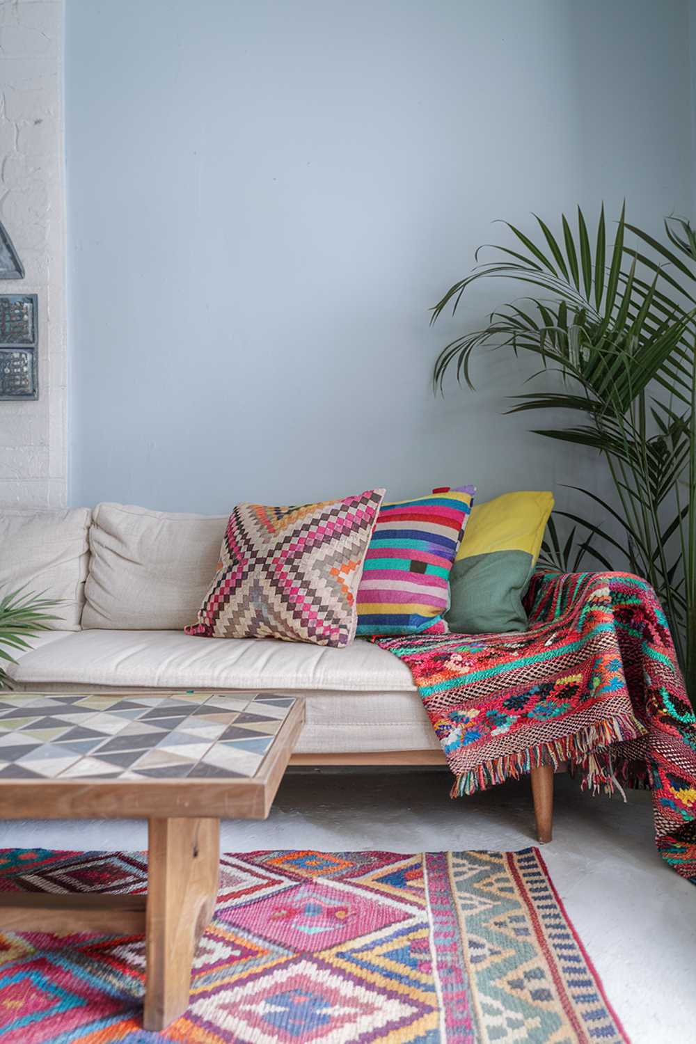 A colorful Scandinavian boho living room decor. There's a beige couch with colorful cushions and a patterned throw. A wooden coffee table with a geometric pattern sits in front of the couch. There's a green plant in the corner. A patterned rug is placed on the floor. The walls are painted light blue.