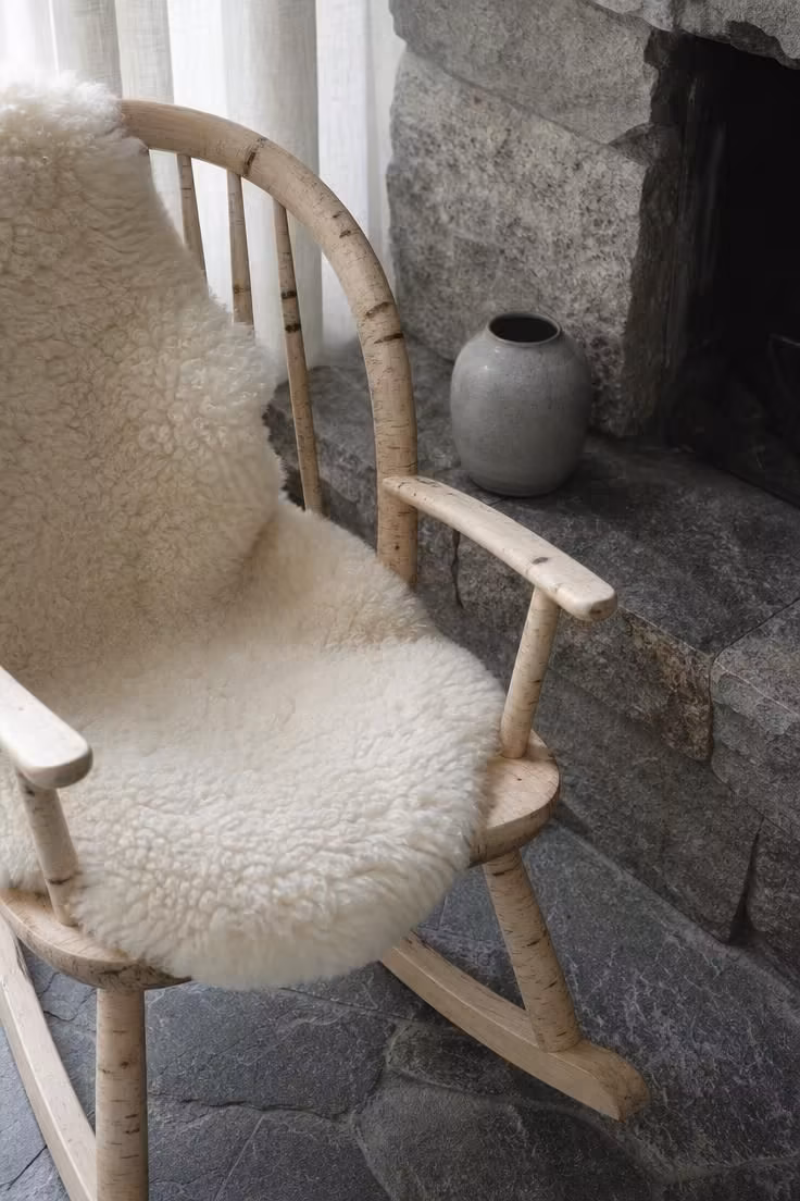Creamy sheepskin draped over a birch rocking chair beside a rough-cut stone hearth with a small ceramic jar in soft diffused light.