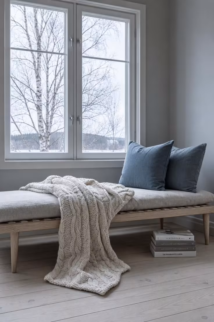 Low-profile ash wood daybed with an oversized cream cable-knit throw and dusty blue linen cushions beneath a large window on an overcast day.