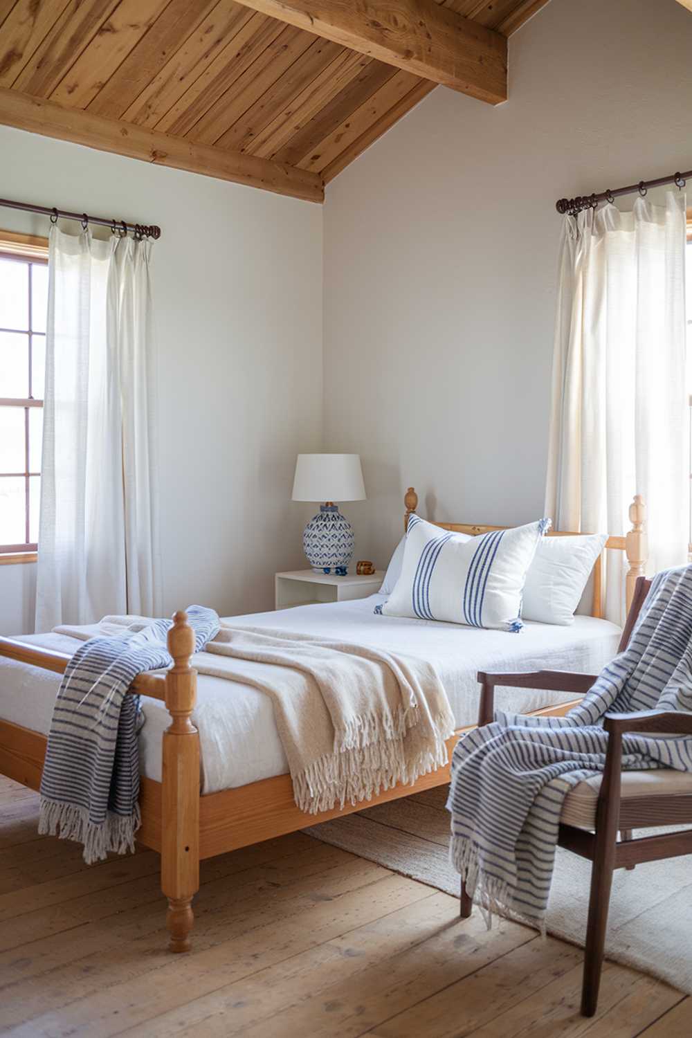 A Scandinavian farmhouse bedroom decor. The room has a wooden beam ceiling, white walls, and wooden floors. There is a wooden bed frame with a white mattress. A beige throw blanket and a white pillow with a blue stripe pattern are placed on the bed. A blue and white striped throw blanket is draped over a wooden chair near the bed. A white lamp with a wooden base is placed on a nightstand beside the bed. The nightstand has a white vase with a blue pattern. The room has a window with white curtains.