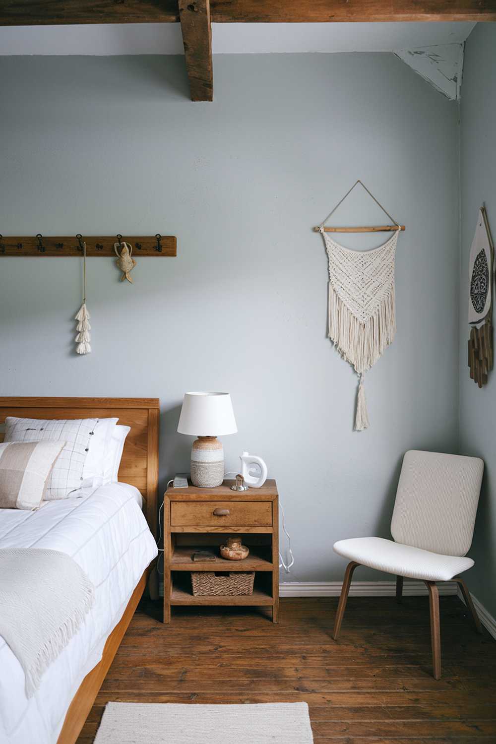 A Scandinavian farmhouse bedroom decor. There's a wooden bed with a white blanket and a few pillows. Beside the bed, there's a nightstand with a lamp and a decorative item. The walls are painted in light blue and have a few hooks and a hanging decoration. The floor is wooden. There's a white chair in the corner. The room has a wooden beam ceiling.