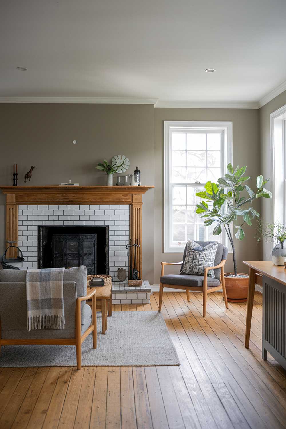 A Scandinavian farmhouse living room. The room has a wooden floor and features a large fireplace with a wooden mantel. There's a cozy sofa with a patterned throw pillow, a wooden chair, and a side table. A potted plant adds a touch of greenery. The walls are painted in a muted gray hue.