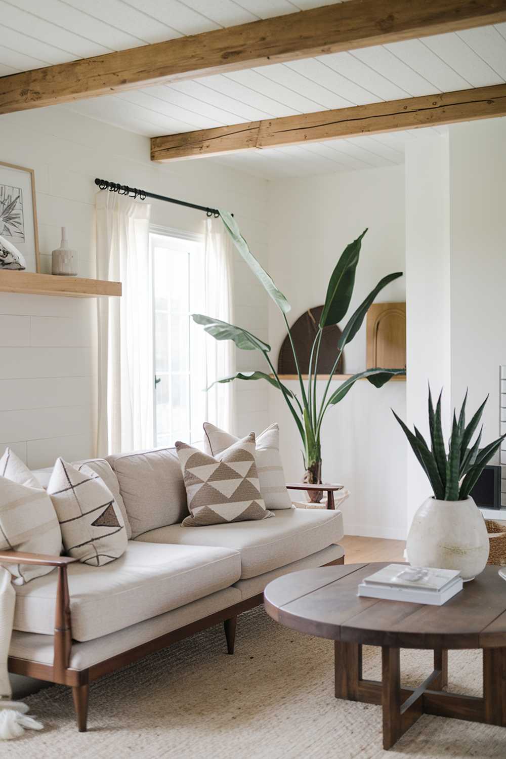 A Scandinavian farmhouse living room design and decor. The room has a beige sofa with wooden legs, a few throw pillows with geometric patterns, a round wooden coffee table, and a large potted plant. The walls are painted white, and there are wooden beams across the ceiling. There is a wooden shelf near the window with a few decorative items. The floor is covered with a beige rug.