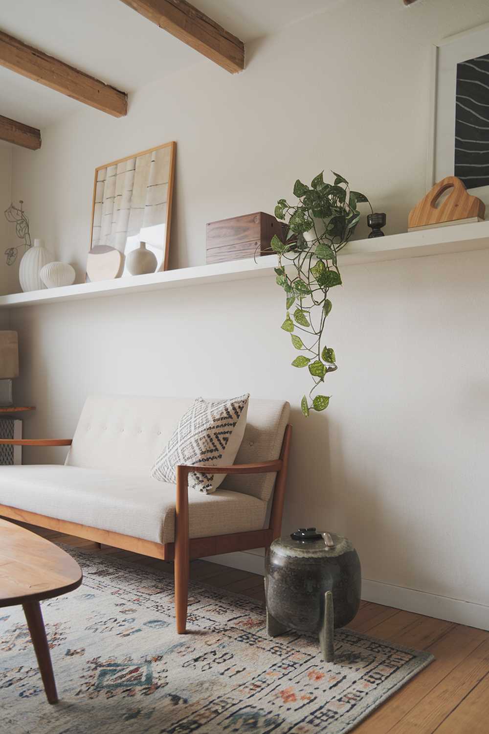 A Scandinavian farmhouse living room. There is a beige sofa with wooden legs and a patterned pillow. Next to the sofa is a wooden coffee table with a round shape and a patterned area rug beneath it. On the wall, there is a white shelf with a few decorative items, including a green plant, a wooden box, and a white vase. The room has a wooden beam ceiling and a few pieces of artwork.