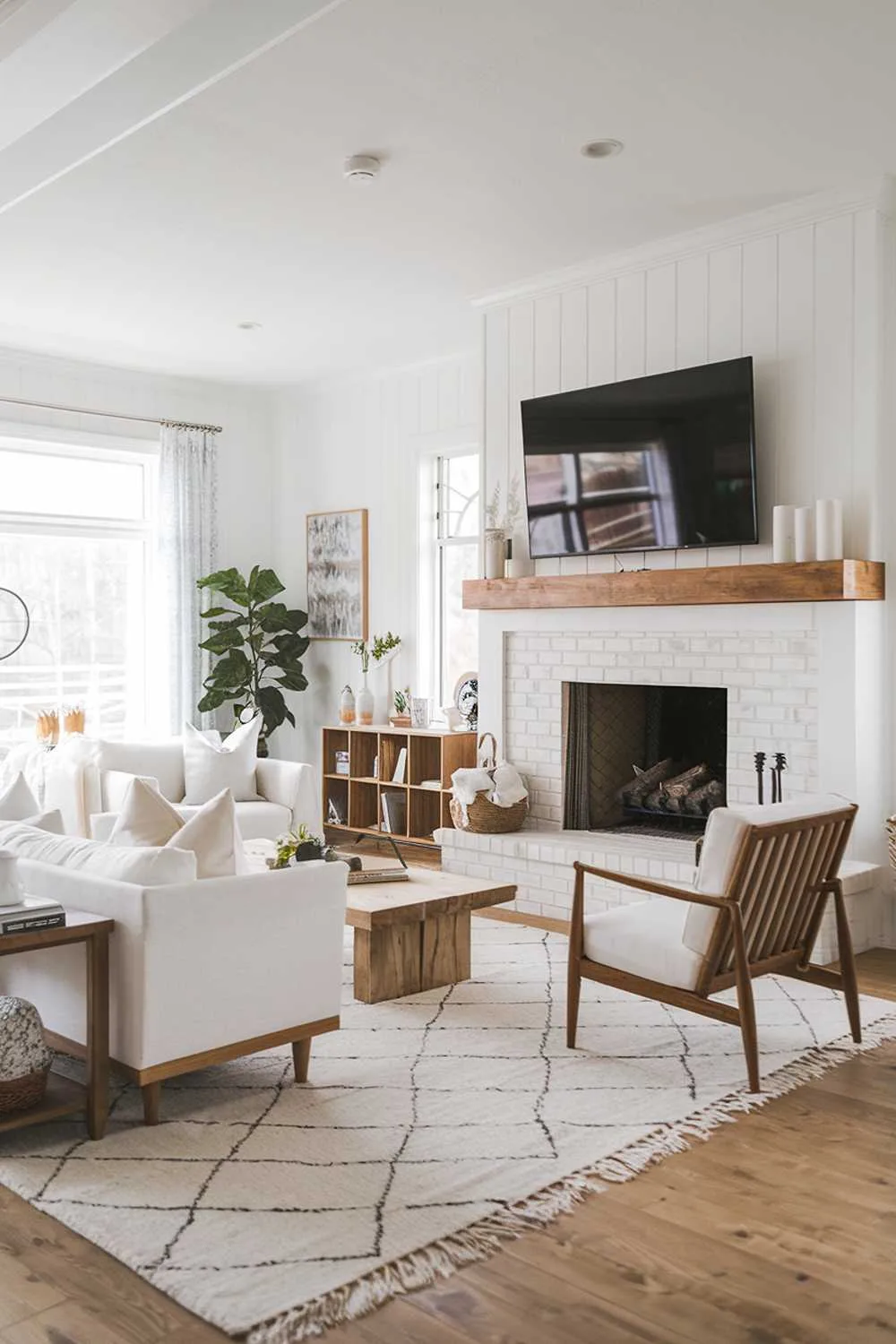 21 Scandinavian Farmhouse Living Room Ideas & Designs 18 A modern Scandinavian farmhouse living room with a white and wood interior. The room has a large fireplace with a wood mantel and a wall-mounted TV. There is a white sofa with wood legs and a wood coffee table in front of the fireplace. A white chair with a wood backrest is placed near the window. The floor is covered with a white wool rug. The walls are adorned with a few pieces of art. A potted plant is placed near the window.