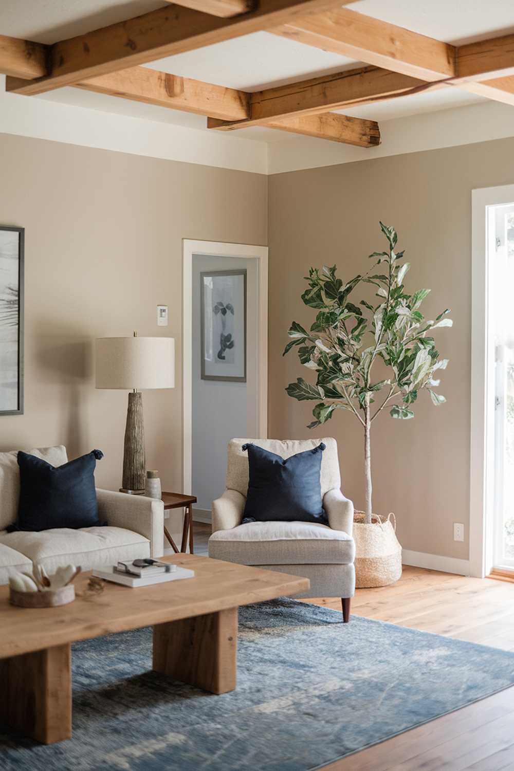 A Scandinavian farmhouse-style living room design and decor. The room has a wooden beam ceiling, beige walls, and a beige sofa with a dark blue throw pillow. There's a wooden coffee table in the middle of the room. A lamp with a beige shade sits on the left side of the room. A potted plant with green leaves is placed on the right side of the room. A blue rug is placed under the coffee table. The floor is made of wood.