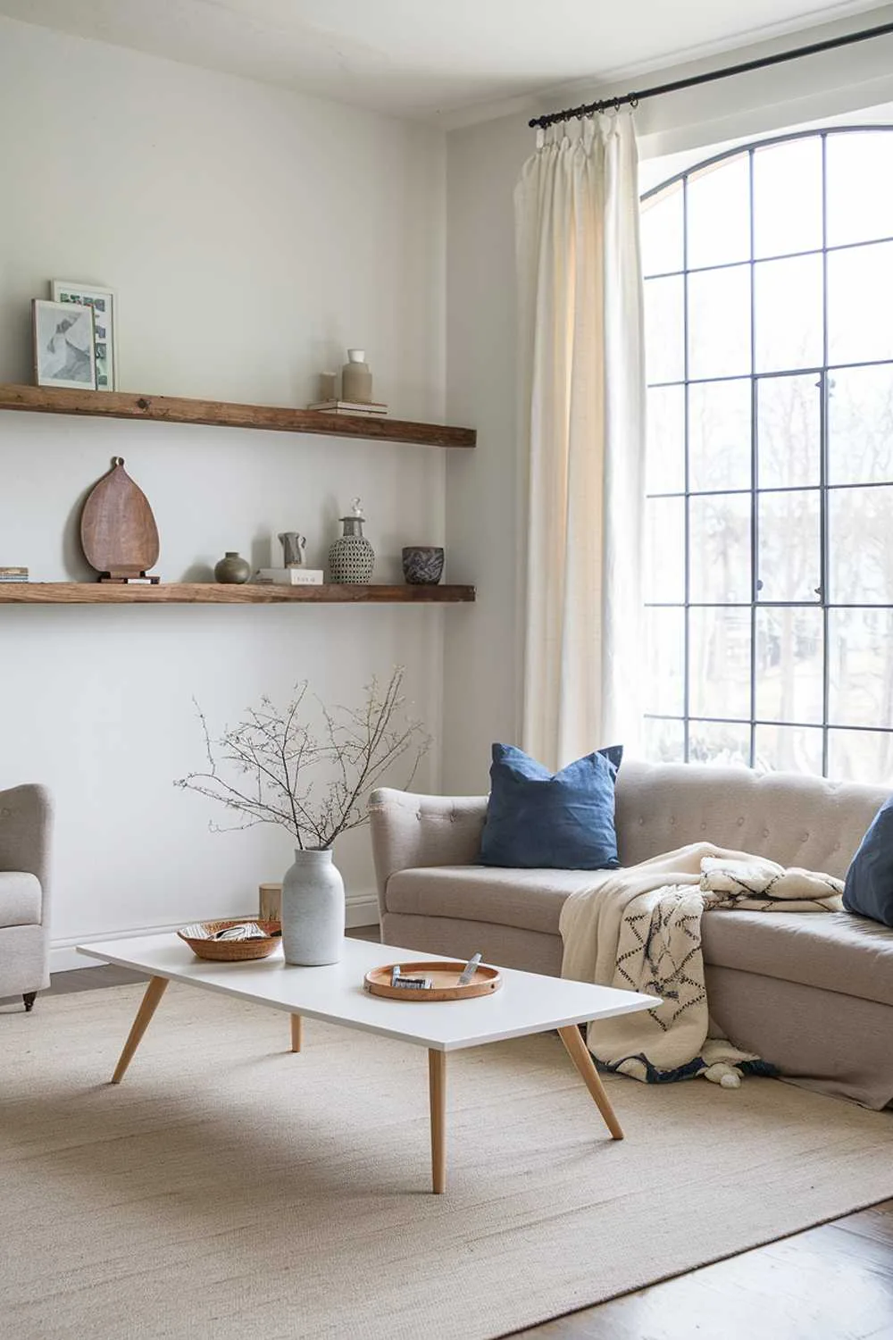 21 Scandinavian Farmhouse Living Room Ideas & Designs 21 A Scandinavian farmhouse-style living room. The room has a beige sofa with blue cushions and a few decorative items on it. There's a white coffee table in the middle of the room. On the left wall, there's a wooden shelf with a few decorative items. The right wall has a large window with a white curtain. The floor is covered with a beige rug.