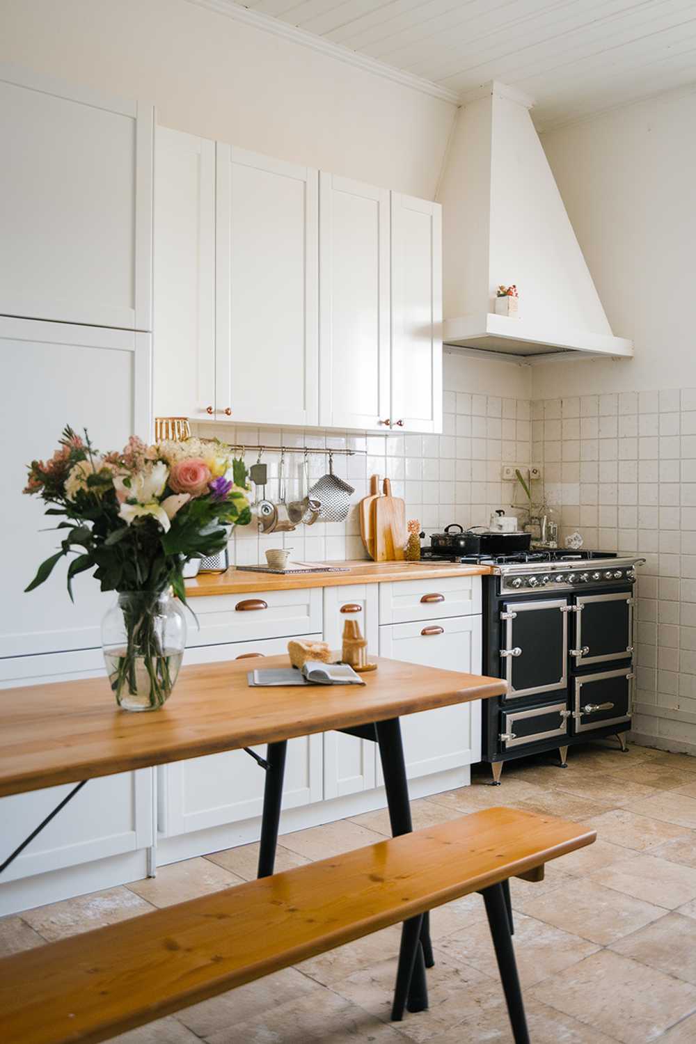 A Scandinavian kitchen design with a wooden table, white cabinets, and a black stove. There is a vase of flowers on the table. The floor is covered with beige tiles.