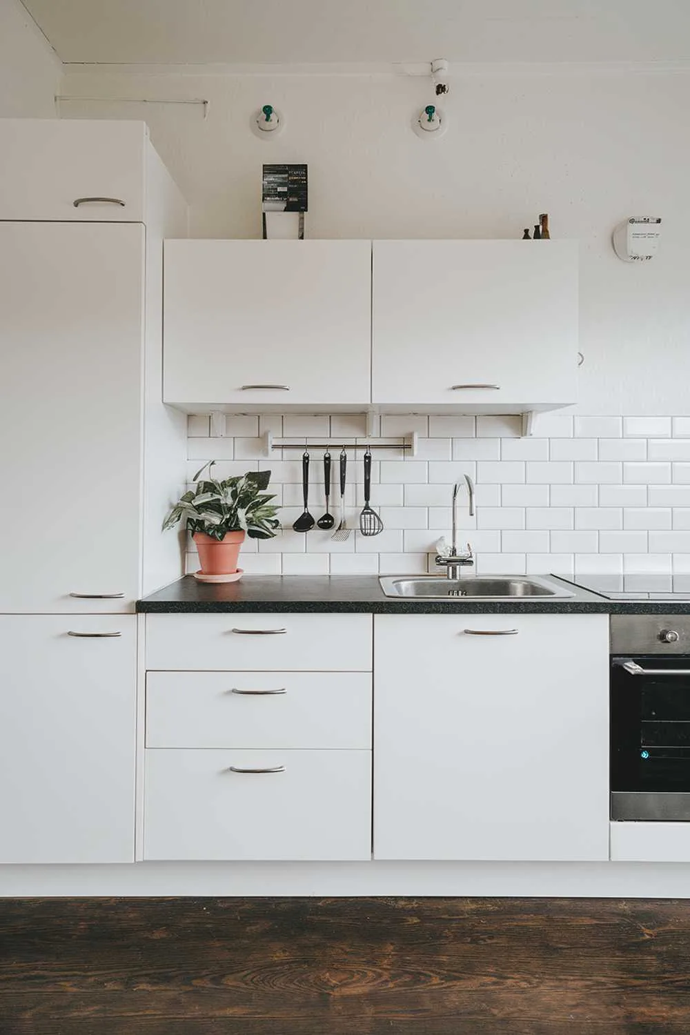 A modern Scandinavian kitchen design with a white cabinet, a black countertop, and a stainless steel sink. There is a potted plant and a few utensils on the counter. The floor is made of dark wood. The wall has a few hooks and a light switch.