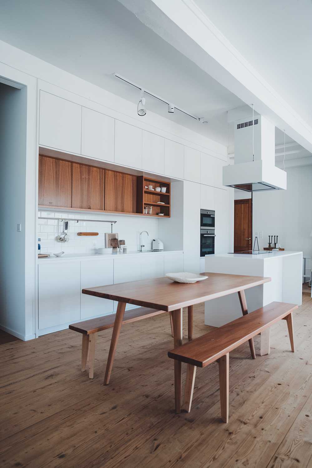 A modern Scandinavian kitchen design. The kitchen has a wooden floor and is painted in white. There is a white cabinet with a wooden front, a white counter, and a white island. There is a wooden table in the middle of the room. The lighting is bright.