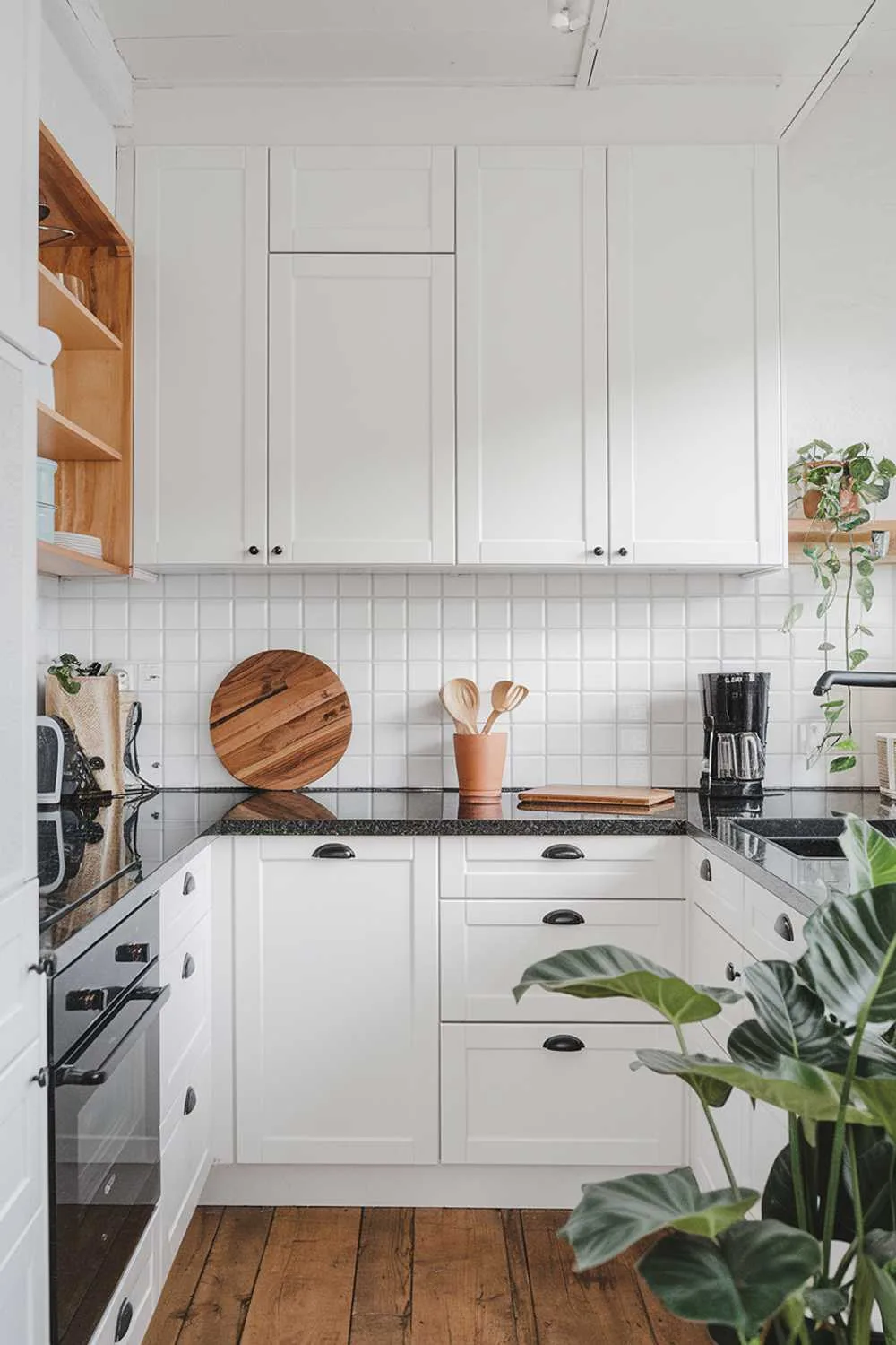 A modern Scandinavian kitchen design with a white cabinets, a dark granite countertop, and a black appliance. The kitchen has wooden elements, including a cutting board and a utensil holder. There is a potted plant near the sink. The floor is made of wooden planks.