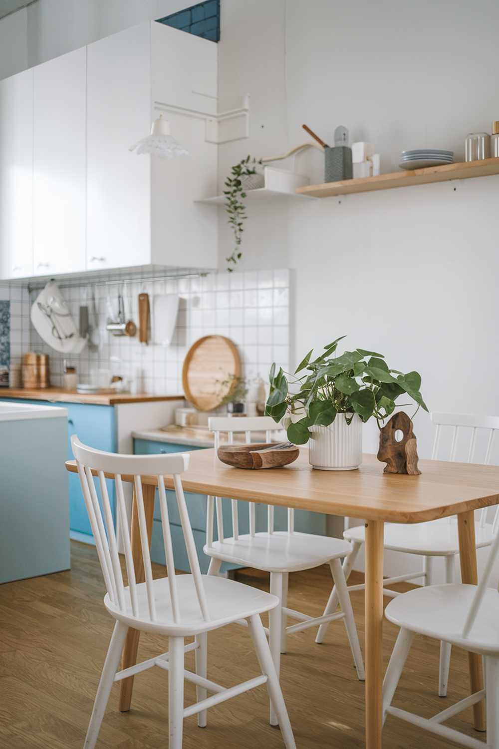 A Scandinavian cozy kitchen design. There's a wooden table in the middle of the room with white chairs around it. On the table, there's a green plant, a white dish with a brown object, and a wooden bowl. In the background, there's a white cabinet, a blue cabinet, and a white wall with a wooden shelf. The floor is made of wood.