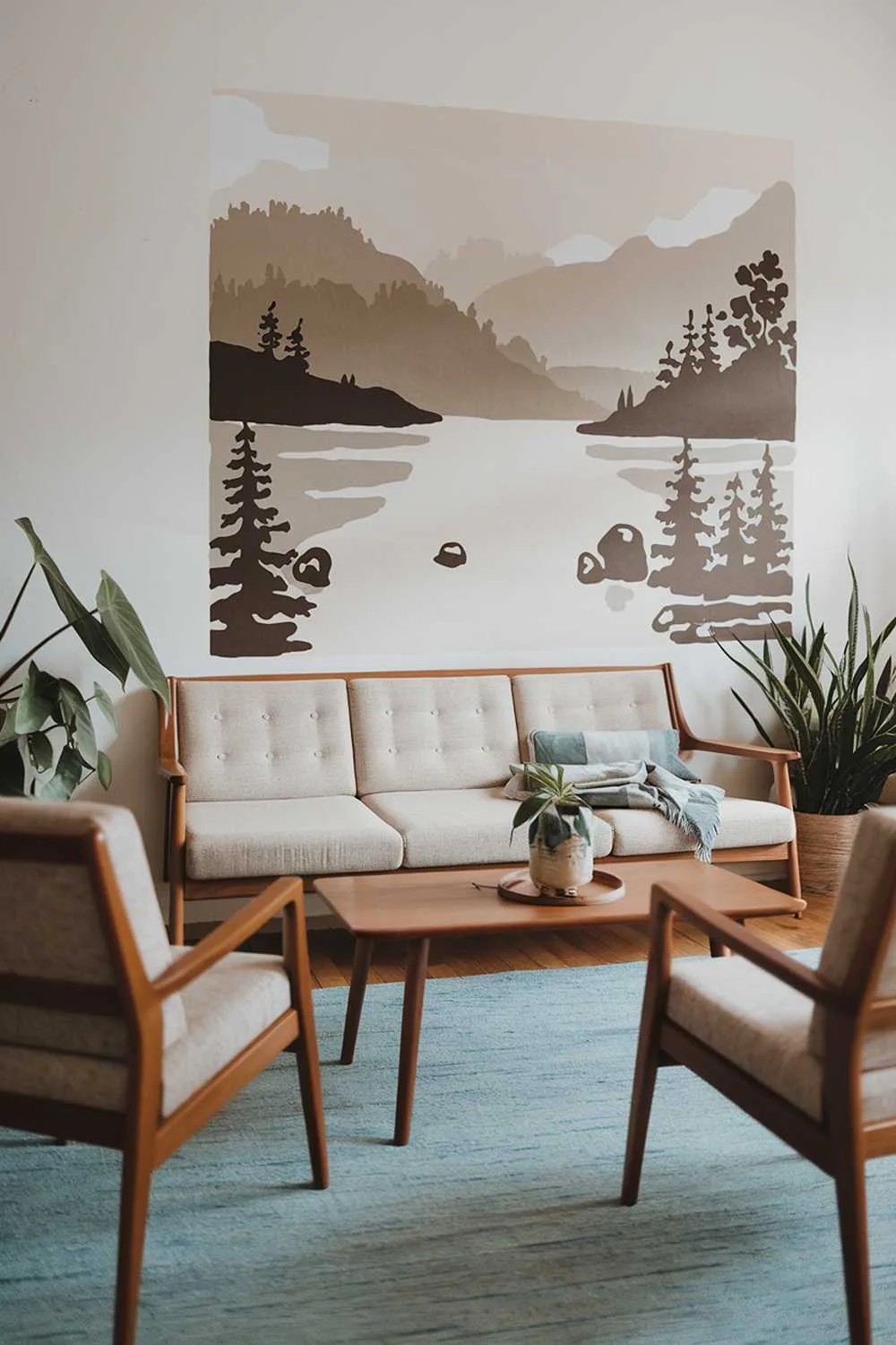 23 Scandinavian Mid-Century Living Room Decor Ideas 11 A Scandinavian modern mid-century living room decor. There's a beige sofa with a wooden frame, a few wooden chairs, and a blue rug. There's a wooden coffee table in the middle. On the wall, there's a large wall art of a serene landscape with mountains, water, and trees. There are a few potted plants in the room. The lighting is soft.