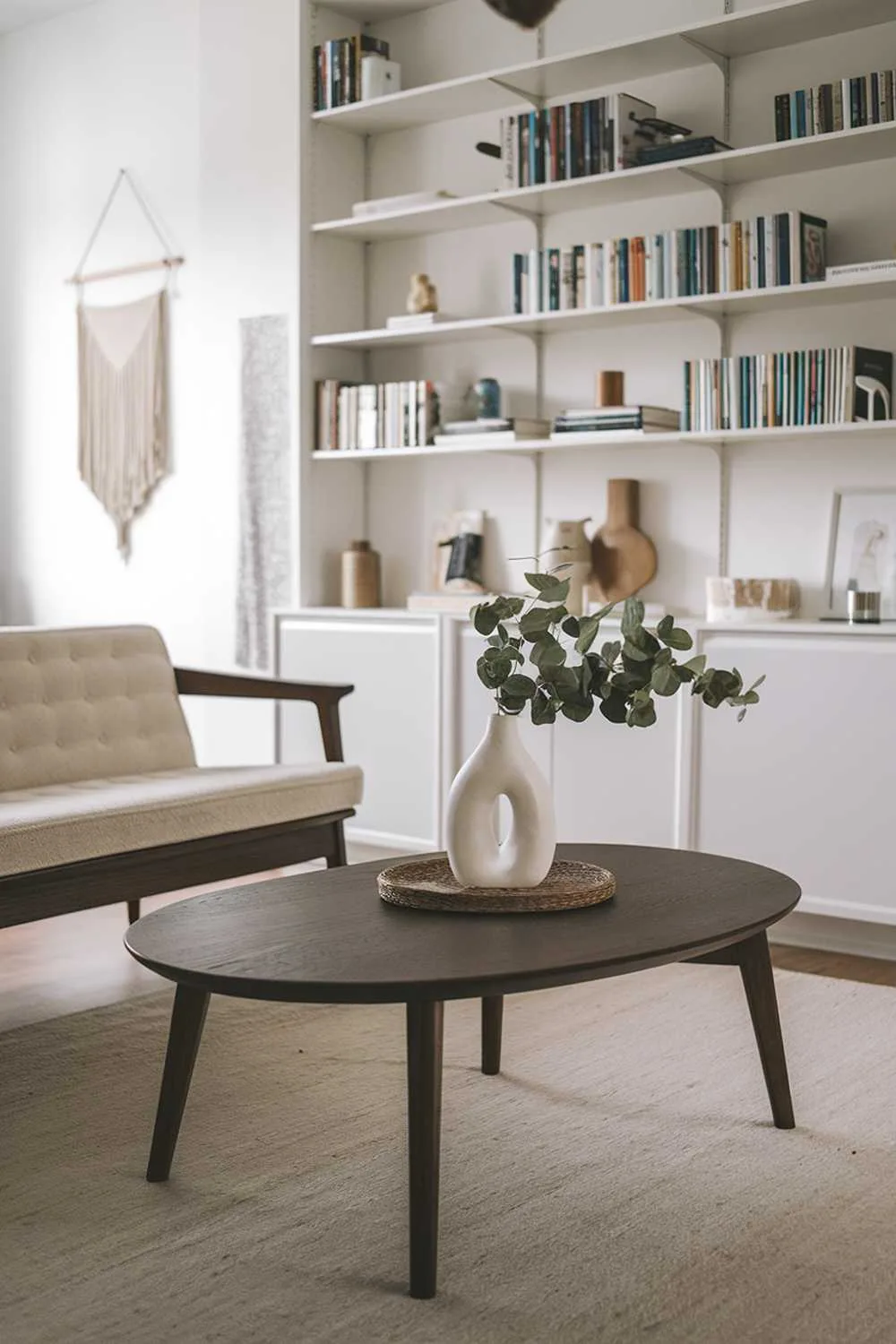 23 Scandinavian Mid-Century Living Room Decor Ideas 23 A mid-century Scandinavian living room decor. The focal point is a dark wooden coffee table with a white vase and a few greenery on top. To the side, there's a beige sofa with a wooden frame. In the background, there's a white bookshelf filled with books and a few decorative items. The floor is covered with a beige rug. The walls have a few hanging items. The overall room has soft lighting.
