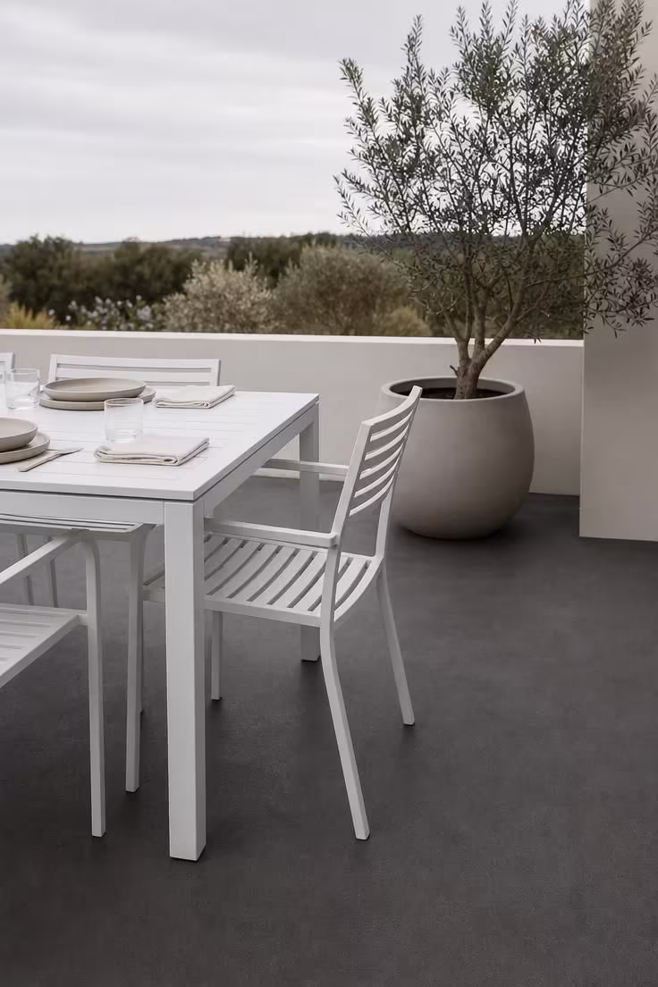 Ash-tinted concrete patio with white powder-coated dining furniture and an olive tree planter under cool morning light