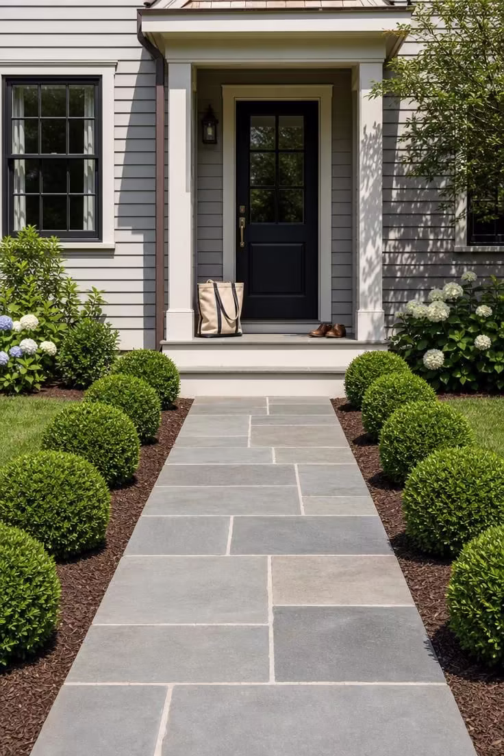 13 Simple Front Yard Landscaping Ideas For Curb Appeal 1 A row of clipped globe boxwoods lining a bluestone front walkway leading to a grey clapboard home.