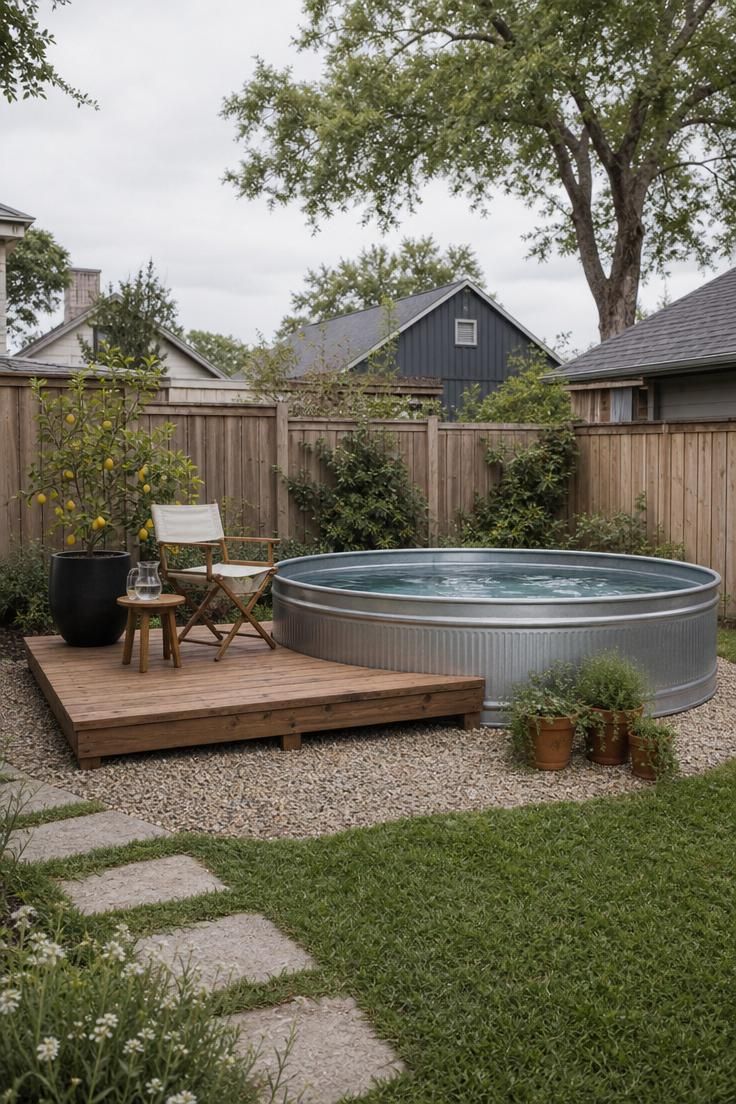 A round galvanized stock tank pool on a pea gravel pad with a cedar deck platform, lemon tree, and folding chair under cool overcast morning light.