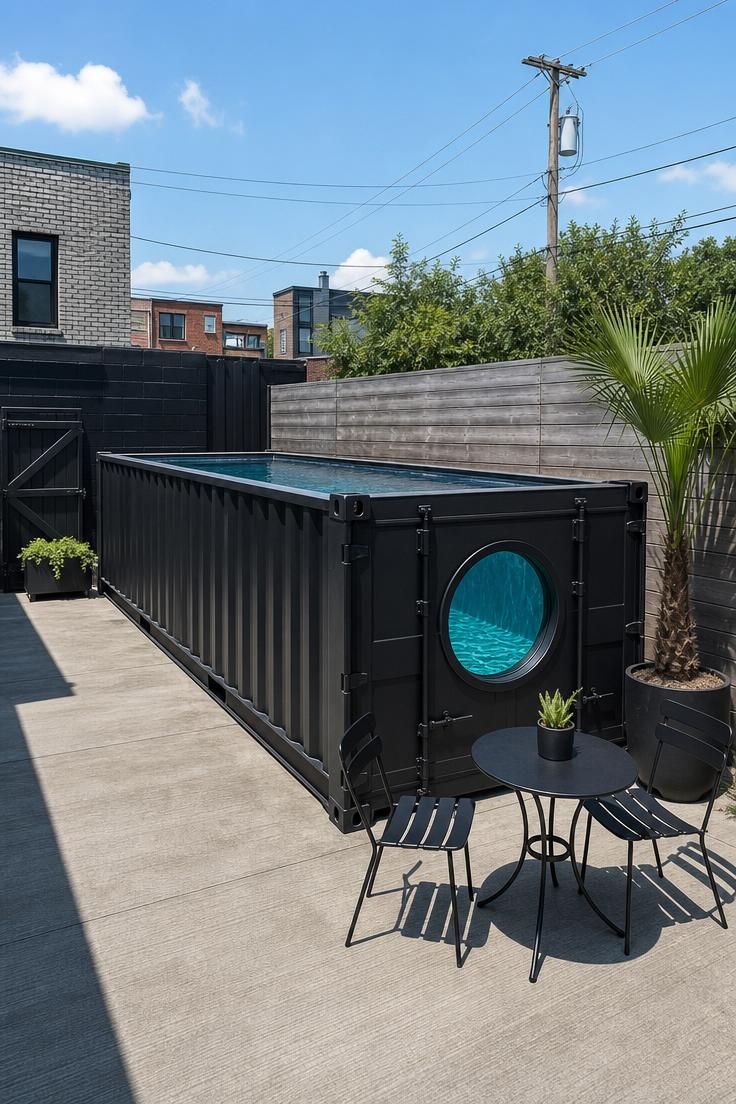 A matte black shipping container pool with a porthole window on a brushed concrete patio under bright midday sun.