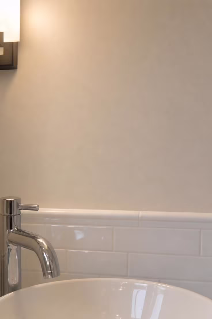 7 Decorating Mistakes That Make a Small Bathroom Without Windows Feel Like a Cave 2 Close-up of a windowless bathroom wall in warm off-white paint, showing how the color reads under soft artificial light next to glossy white tile trim and a polished chrome faucet.