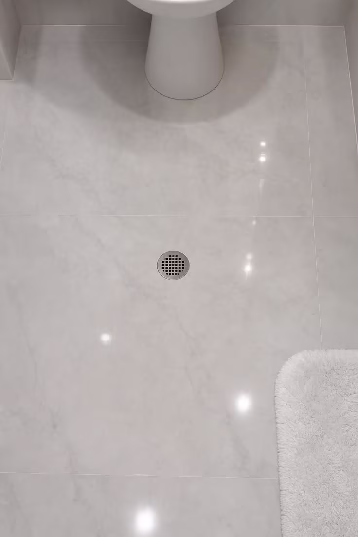 7 Decorating Mistakes That Make a Small Bathroom Without Windows Feel Like a Cave 4 Overhead view of glossy large-format pale porcelain tile on a windowless bathroom floor, showing reflected light moving across the polished surface with thin grout lines barely visible.