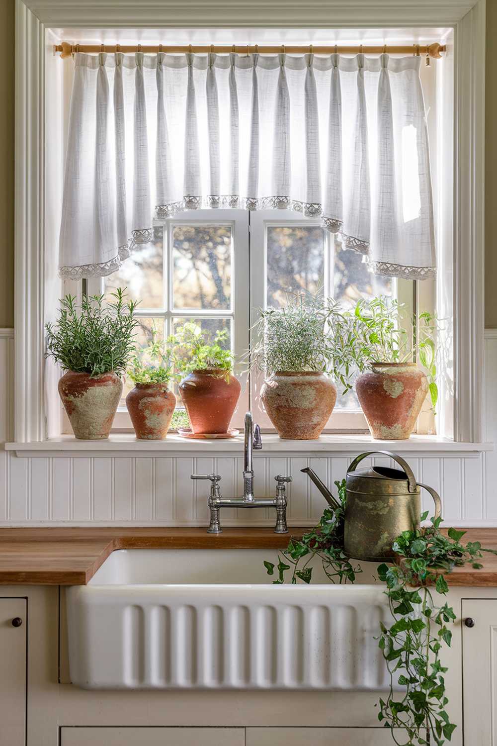 A sunny kitchen window dressed with white linen cafe curtains gently filtering light onto a farmhouse sink below. The deep windowsill holds a collection of terracotta pots filled with fresh herbs and a small vintage watering can. Below, white beadboard adds texture to the wall, while a vintage-inspired faucet in aged brass completes the scene.