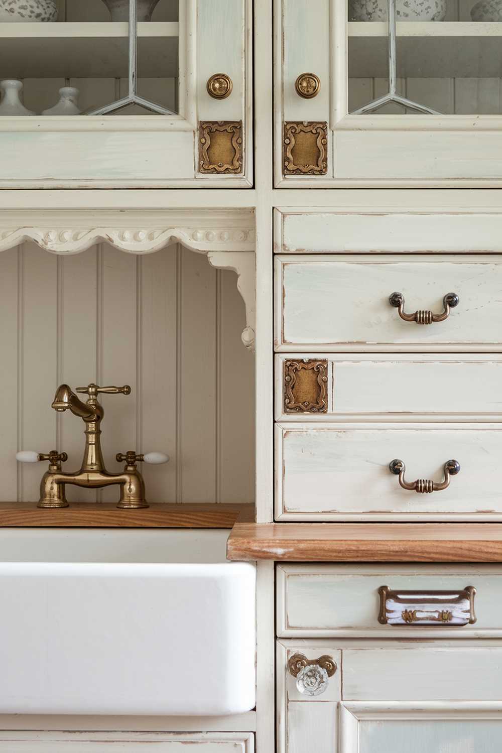 Close-up details of a cottage kitchen showing vintage-inspired brass bin pulls and glass knobs on pale blue cabinet doors. Decorative hinges add character, while a Victorian-style tap in aged brass stands out against a white farmhouse sink. Beadboard backing and delicate trim work complete the authentic cottage look.