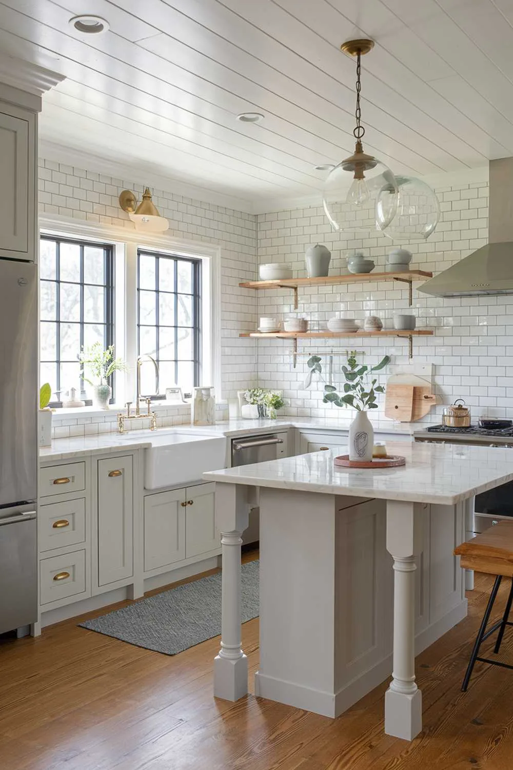 A bright, updated cottage kitchen featuring pale gray cabinets with modern hardware, balanced by traditional elements like a farmhouse sink and subway tile backsplash. Contemporary pendant lights with clear glass shades hang from a shiplap ceiling, while a compact island with a marble top provides a sleek workspace. Stainless steel appliances blend seamlessly with vintage-inspired details.