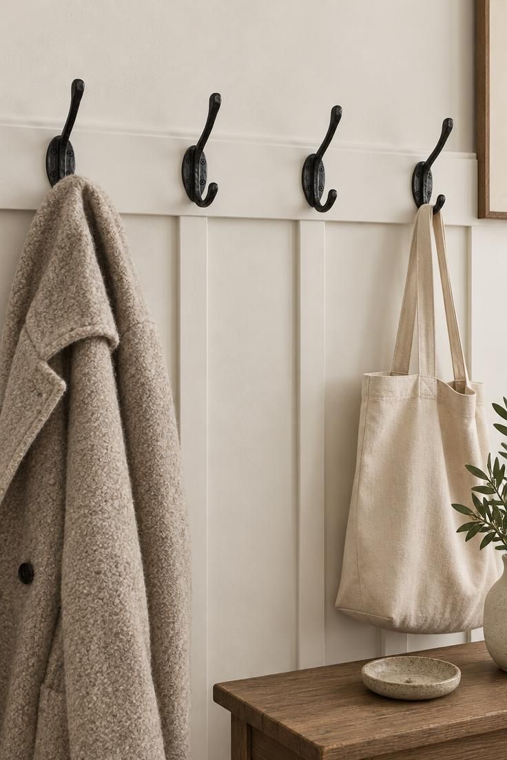 13 Cozy Small Entryway Ideas That Make Coming Home Calm 12 A white board-and-batten rail across a wall, with four black wrought-iron hooks holding a wool coat, a canvas tote, and two empty hooks, above a narrow wood console.