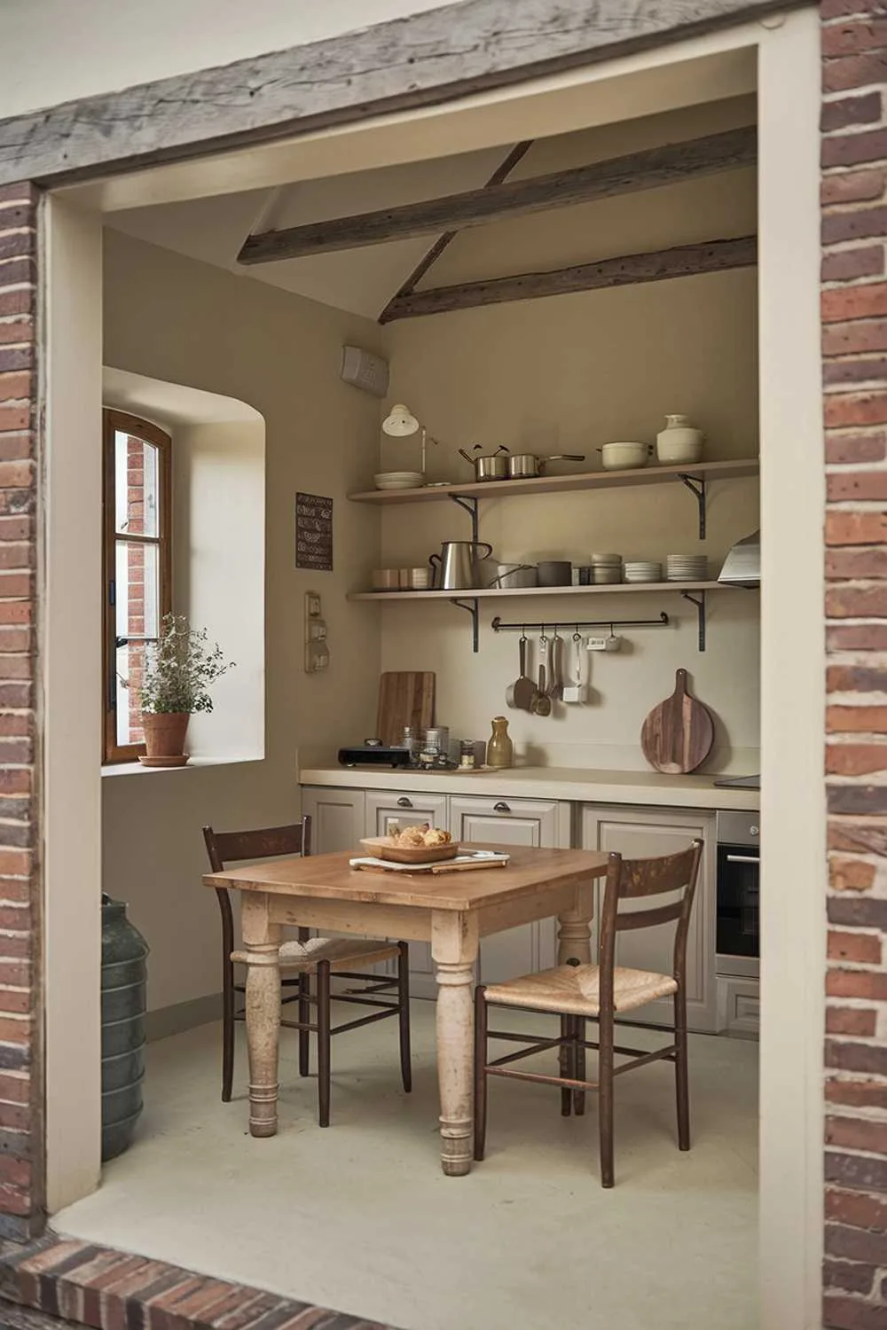 21 Small Farmhouse Kitchen Ideas for Country Style Comfort 19 A small farmhouse kitchen interior design. There is a wooden table in the center of the room with a few chairs. The walls are painted beige, and there are a few shelves with cookware and utensils. There is a window near the entrance with a flower pot. The room has a rustic charm with its exposed brick wall and wooden beams.
