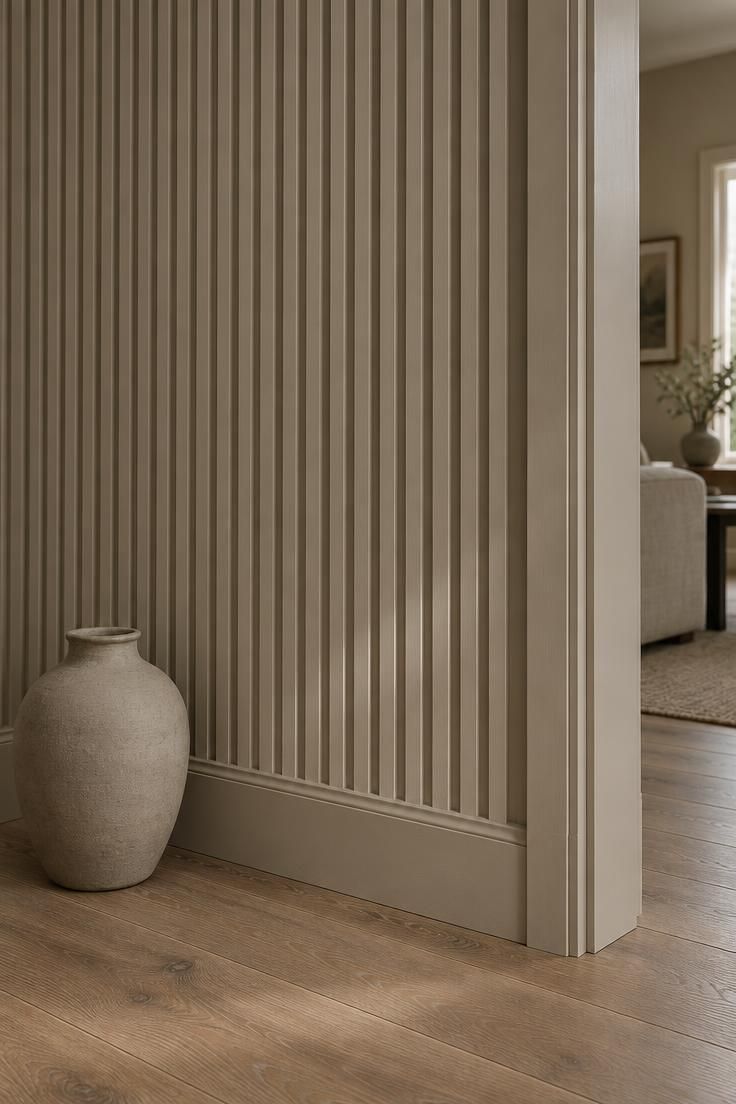 A hallway wall clad in warm putty vertical fluted wood panels from baseboard to ceiling, meeting a bare wood floor beside a simple door frame.
