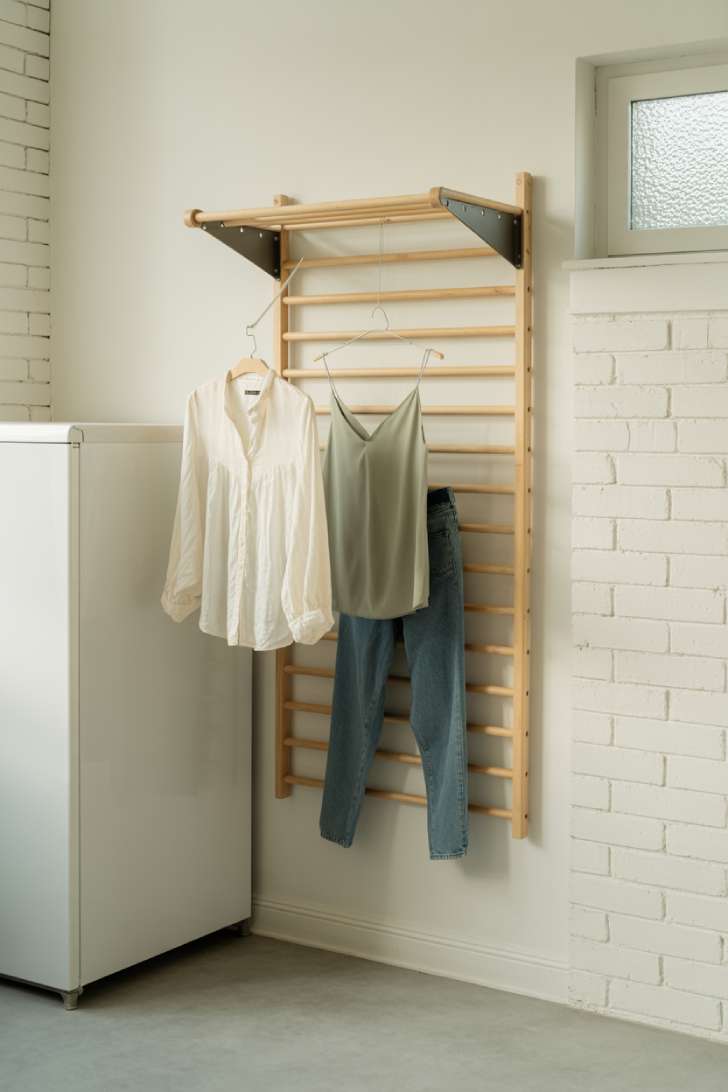 Wall-mounted wooden fold-flat drying rack extended with delicate garments hanging in a white laundry room