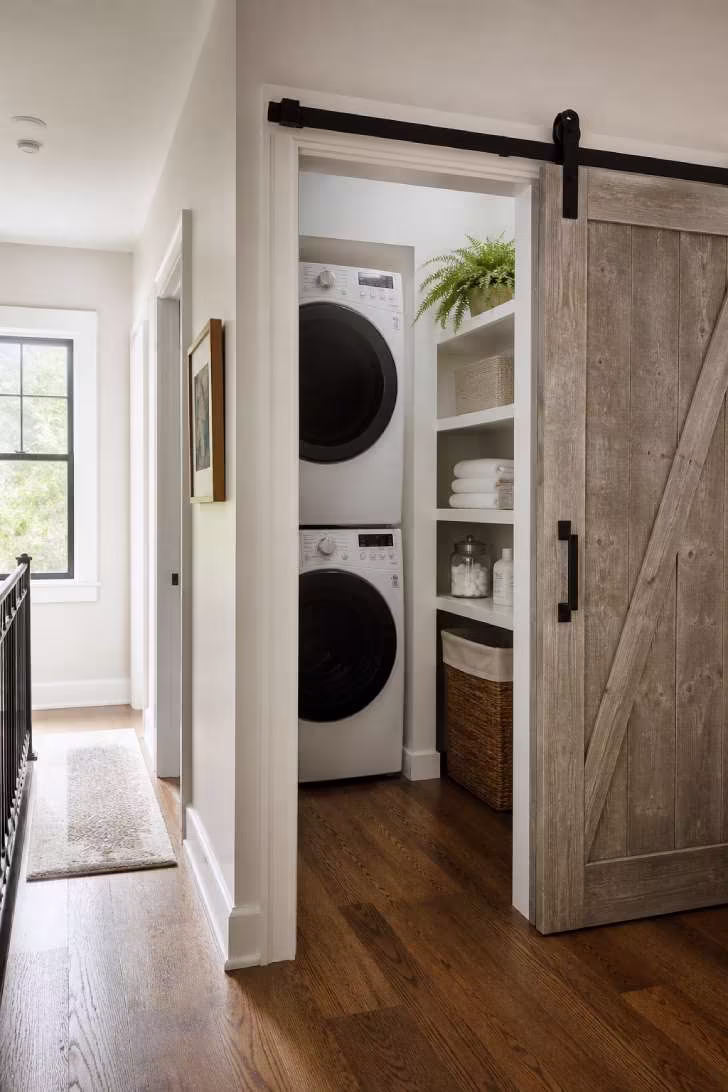 A gray-washed sliding barn door opened to reveal a compact organized laundry room with stacked machines and open shelving