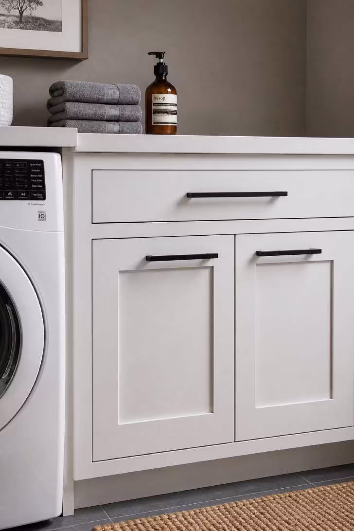 Close-up of matte black bar pulls on white shaker cabinets in a small laundry room with an amber glass soap dispenser