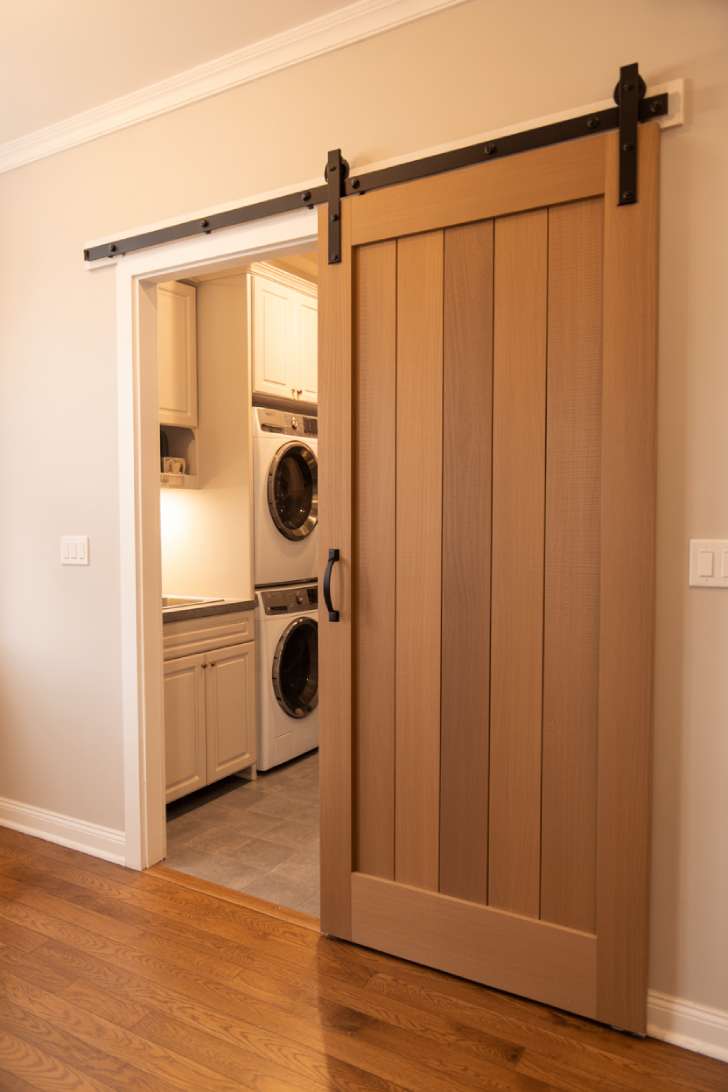 A natural wood sliding barn door on a black rail half-open to a narrow laundry room with white cabinets inside