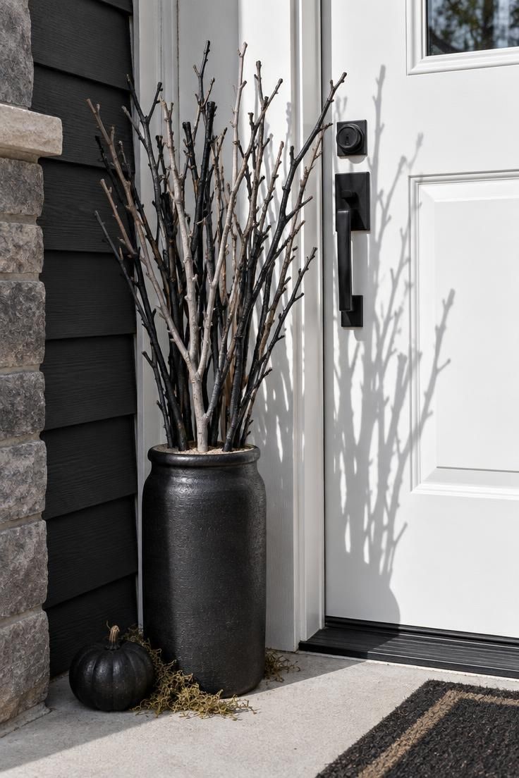 A matte black stoneware crock filled with painted black and charcoal branches standing beside a white front door on a pale concrete stoop.