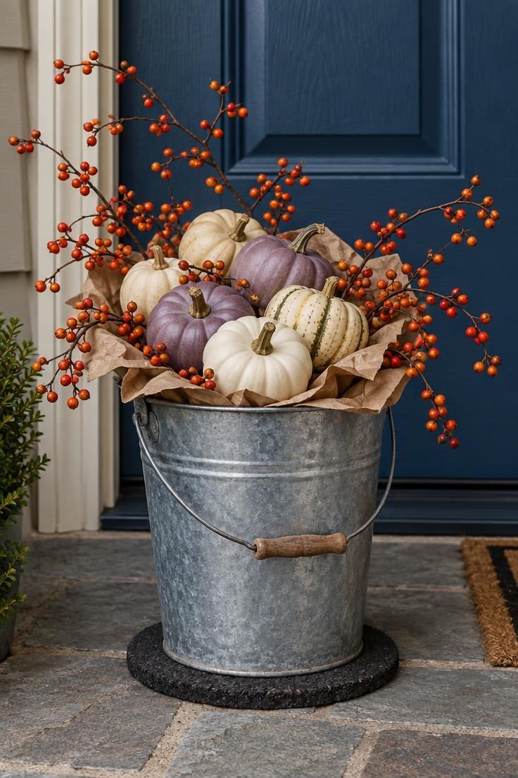 A galvanized pail filled with cream, white, and muted purple mini pumpkins plus red-orange bittersweet branches against a deep blue door.