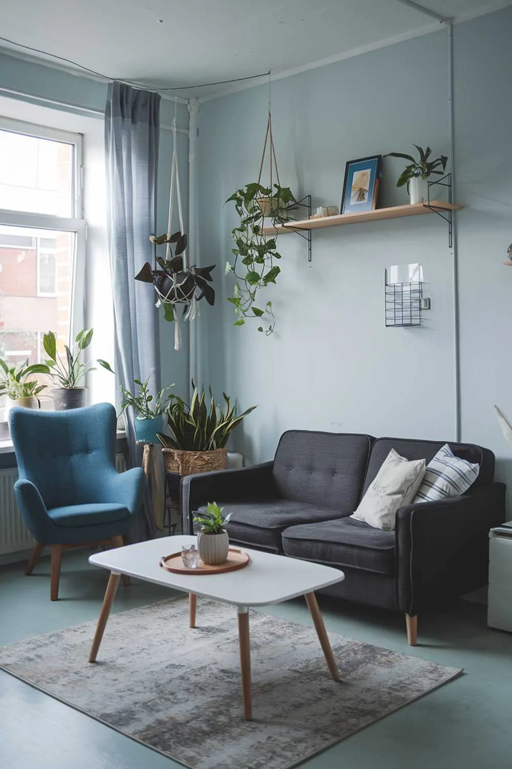 A cozy small Scandinavian living room with a dark gray sofa and a white coffee table in the middle of the room. There's a blue chair next to the window. A wooden shelf is placed above the sofa, holding a few decorative items. The walls are painted in light blue and have a few hanging plants. A rug is placed on the floor.