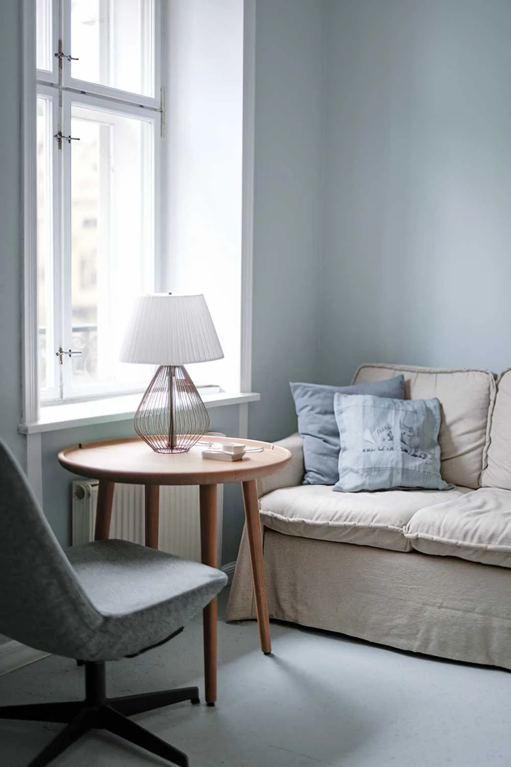 A cozy Scandinavian living room with a small, round wooden table near the window. There's a white lampshade on the table. A beige sofa with a few cushions is placed against the wall. A gray chair with a wooden base is placed near the sofa. The walls are painted in light blue.