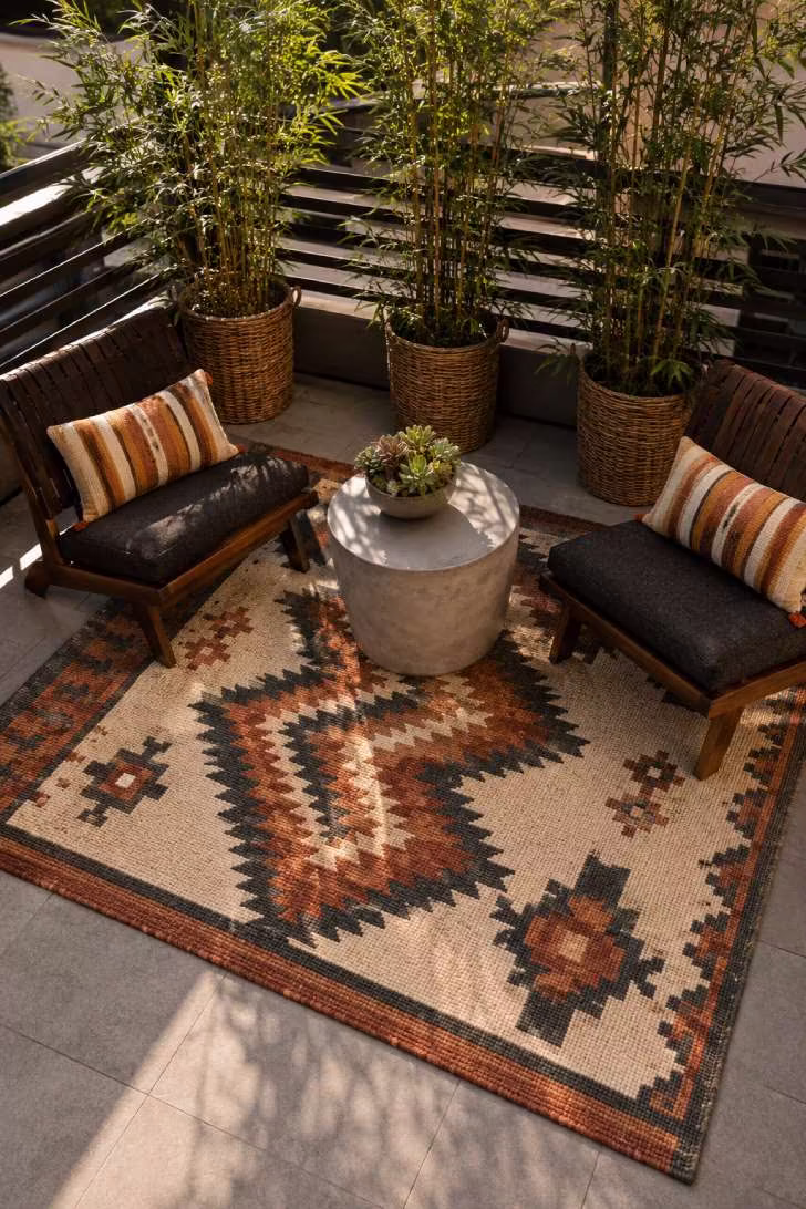 Small terrace with geometric outdoor rug in terracotta and cream defining a lounge zone with low wooden chairs and potted bamboo
