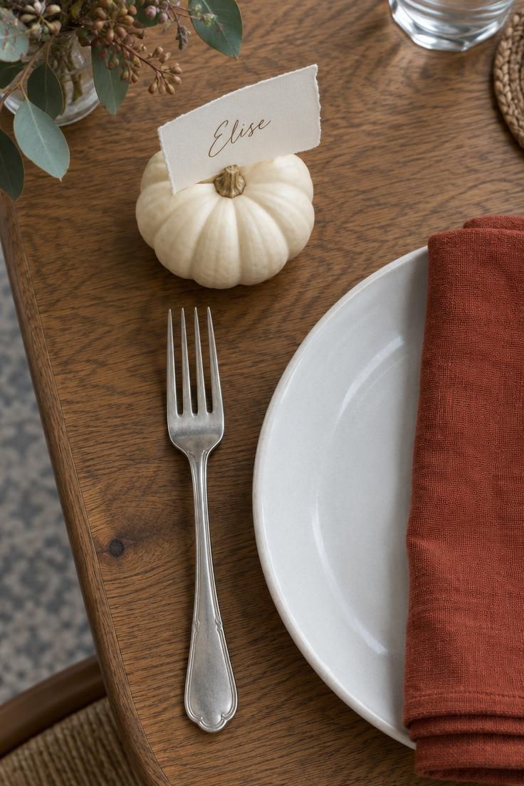 10 Warm Small Thanksgiving Table Decor Ideas for Close Meals 7 An overhead view of one place setting with a white mini pumpkin holding a handwritten torn-edge name card, a rust napkin, a white plate, and a silver fork.