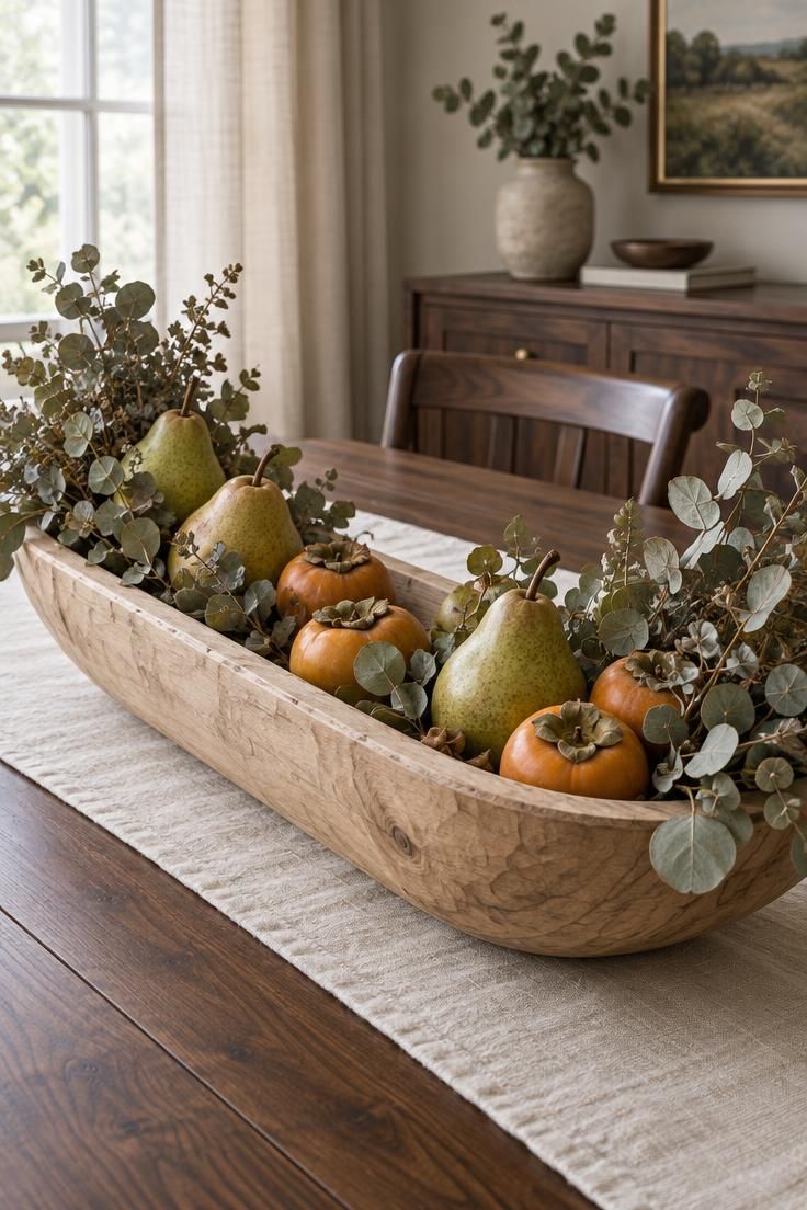 10 Warm Small Thanksgiving Table Decor Ideas for Close Meals 8 A close-up of a light wood dough bowl on a dark walnut table filled with pears, persimmons, and dried eucalyptus, with a cream linen runner behind.