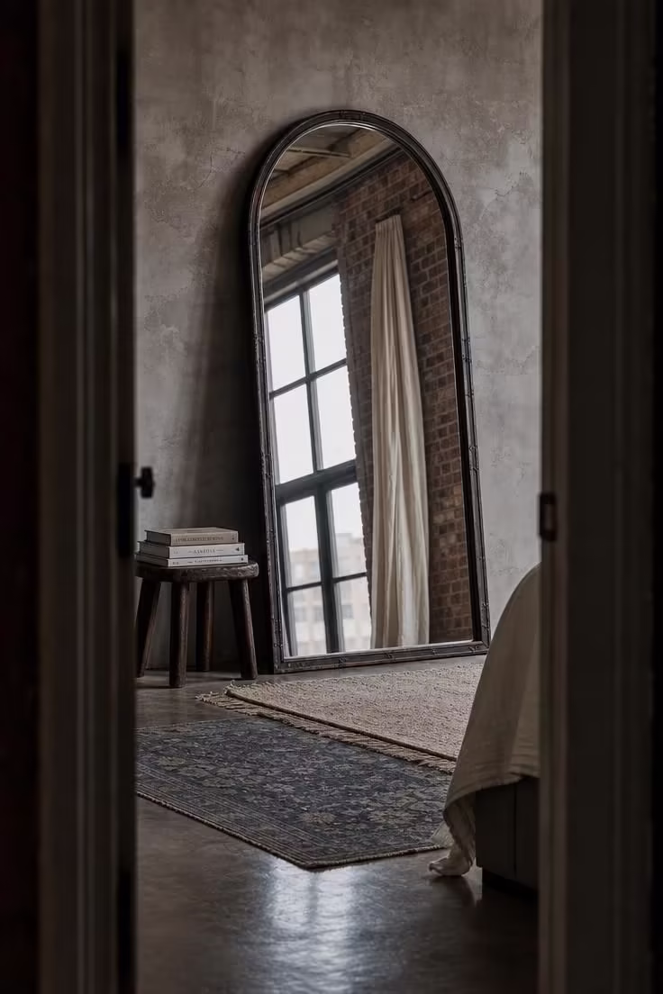 Large aged iron arch mirror leaning against a cement wall with layered rugs on concrete floors