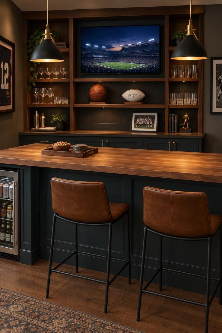 11 Cozy Sports Man Cave Ideas for Game-Day Comfort 5 A butcher-block bar counter with two leather stools, open walnut back-bar shelving with glasses and signed balls, a screen above, brass pendants over each end, and a beverage fridge in the base under warm light.